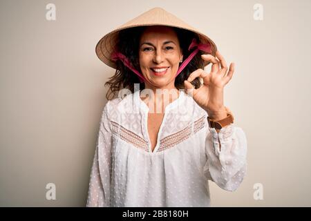 Donna brunetta di mezza età che indossa il cappello conico tradizionale asiatico su sfondo bianco sorridendo positivo facendo il segno ok con la mano e le dita. Riuscito Foto Stock