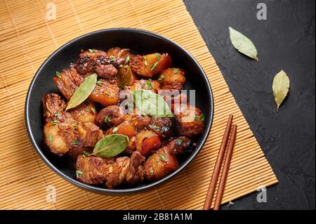 Adobo maiale in ciotola nera su sfondo ardesia scuro. Maiale Adobo o Adobong Baboy è un piatto di cucina filippina con pancia di maiale brasata, foglie di alloro, salsa di soia Foto Stock
