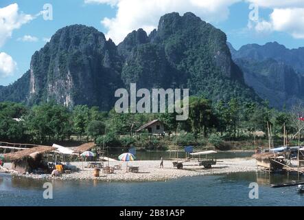 Caffè e mercato realizzati su una sponda di ciottoli nel mezzo del fiume Nam Song a Vang Vien (Vang Vieng), nella provincia di Vientiane, nel Laos. I tetti di paglia coprono le bancarelle e gli ombrelloni multicolore. Una tradizionale casa in legno del Laos si trova sulla riva lontana con alberi coperti, montagne carsiche alle spalle. Foto Stock