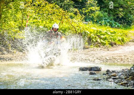 Estate giornata di sole nella foresta. L'atleta ENDURO supera un ruscello poco profondo con molti spruzzi d'acqua Foto Stock