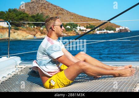 Felice giovane uomo si sente felice sulla barca a vela di lusso catamarano in mare turchese nelle vacanze estive sull'isola. Modello maschio caucasico. Foto Stock