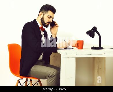 Uomo barbuto, barba corta. Responsabile serio caucasico con baffi scrivere a blocco note, parlando al telefono cellulare al tavolo di legno bianco indossando giacca nera, camicia bianca, cravatta a prua rossa isolato su bianco Foto Stock