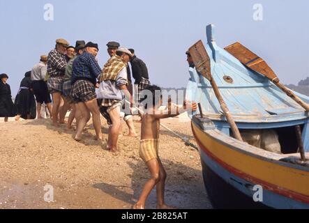 Nazare, Portogallo-12 luglio 1980: Pescatori e ragazzino che tira una tipica vecchia barca portoghese a riva sulla spiaggia di Nazare, Portogallo. Foto Stock