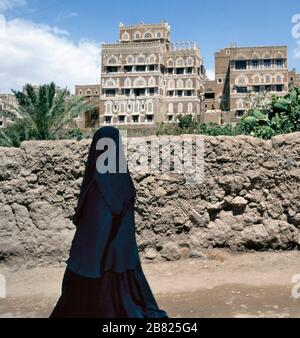 Una donna musulmana velata passeggiate su una sana di una strada, Yemen.a sfondo tipiche case dello Yemen. Foto Stock