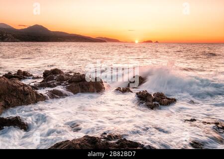 Onde che si schiantano sulla costa rocciosa della Corsica mentre il sole tramonta sulla città Balagne di Ile Rousse Foto Stock