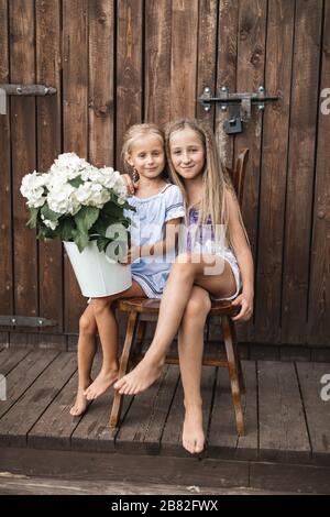 Due ragazze bionde felici capelli, sorelle o amici, seduti insieme sulla sedia di legno vicino al vecchio fienile di legno all'aperto, e tenendo fiori bouquet, w Foto Stock