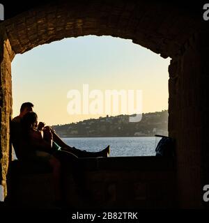 Giovane coppia innamorata gode della calda luce del tramonto che si siede abbracciandosi l'un l'altro di fronte alla bellezza del mare napoletano incorniciato da un vecchio arco in pietra Foto Stock