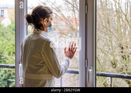 Un infermiere che indossa una maschera medica per il viso e un cappotto bianco, è in piedi inattivo davanti a una finestra chiusa, fissandosi nello spazio. Foto Stock