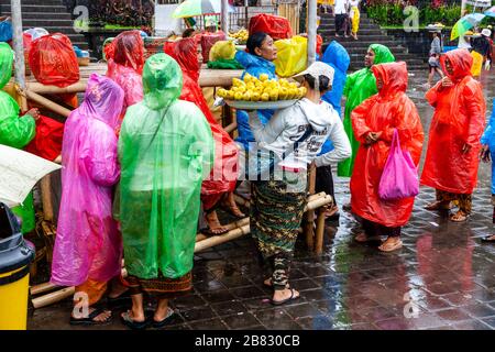 Donne locali che vendono mais/mais ai visitatori che indossano Ponchos durante UNA doccia a pioggia, la cerimonia di Batara Turun Kabeh, il tempio di Besakih, Bali, Indonesia. Foto Stock