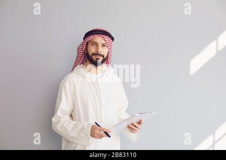 Attraente uomo arabo sorridente scrive in appunti su sfondo grigio Foto Stock