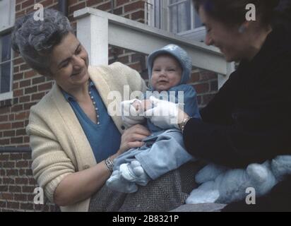 Due donne mature tengono un bambino vestito in un vestito blu fuori di una casa suburbana, 1965. () Foto Stock