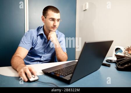 Giovane poliziotto in uniforme e occhiali seduti in ufficio e che lavorano al laptop su pareti di sfondo. Poliziotti in azione concetto Foto Stock