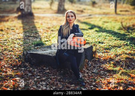 bella giovane donna bionda con stravagante make-up in una giacca di pelle nera con ampi occhi aperti e una bocca aperta con una zucca in mano Foto Stock