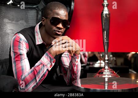 Giovane uomo africano bello in elegante abbigliamento casual e occhiali da sole seduta in grande poltrona in pelle nera nel ristorante su sfondo parete rosso. Tre Foto Stock