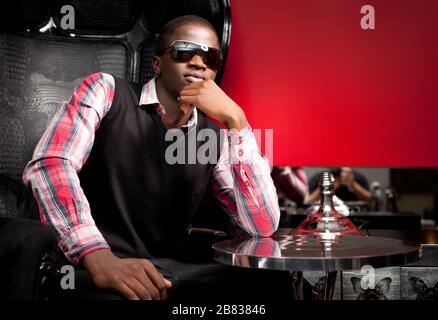 Giovane uomo africano bello in elegante abbigliamento casual e occhiali da sole seduta in grande poltrona in pelle nera nel ristorante su sfondo parete rosso. Tre Foto Stock