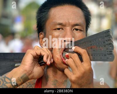 Il devoto taoista cinese thailandese (mAh song) spierce la sua guancia destra con i governanti del metallo durante il Festival vegetariano di Phuket (Nine Emperor Gods Festival). Foto Stock