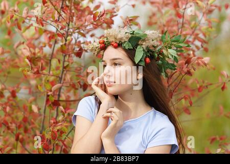 Bella ragazza dai capelli lunghi in una t-shirt e una corona di fiori e bacche. Ritratto autunnale su sfondo di foglie rosse Foto Stock