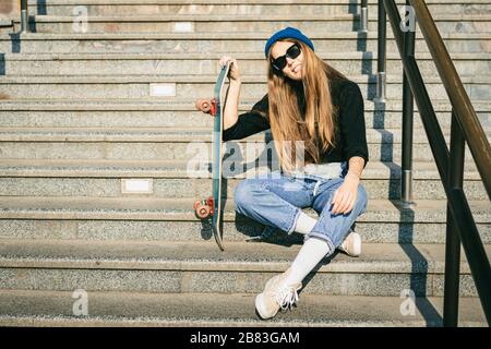 Donna elegantemente vestita con jumpsuit in denim blu con skateboard. Foto della strada. Ritratto di ragazza con skateboard. Stile di vita, concetto giovanile Foto Stock