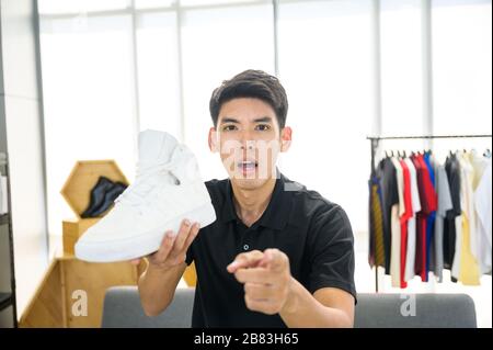 Social media influencer che rivede la moda scarpe. Sorridente giovane uomo che vlogging circa la scarpa sportiva degli uomini e filmarsi a casa su una videocamera. Foto Stock