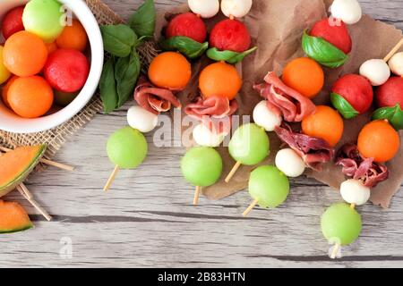 Spiedini di frutta estiva colorati con melone, formaggio e prosciutto, primo piano su uno sfondo di legno Foto Stock