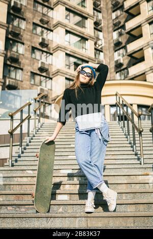 Donna elegantemente vestita con jumpsuit in denim blu con skateboard. Foto della strada. Ritratto di ragazza con skateboard. Stile di vita, concetto giovanile Foto Stock