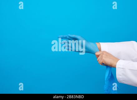 medico femminile in un cappotto bianco mette sulle sue mani i guanti medici sterili del lattice blu sulle sue mani, sfondo blu, posto per il testo Foto Stock