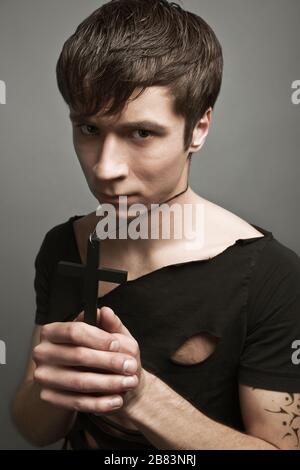 Giovane uomo in moderno abbigliamento nero e libertà tatuaggio tenendo grande croce nera in mano con gli occhi chiusi. Foto Stock