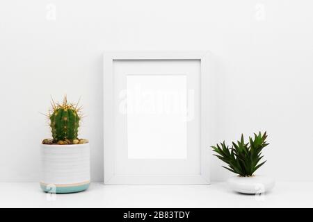 Mokup cornice bianca con cactus e succulente su uno scaffale o scrivania. Schema colore bianco. Orientamento verticale della cornice. Foto Stock