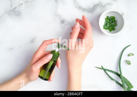 Donna che applica crema idratante organica per le mani. Bottiglia di lozione Aloe vera in mani femminili e steli di piante affettati su fondo marmoreo. Dermatologia delle mani Foto Stock