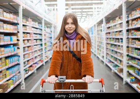 Giovane donna con carrello di alimentari e scaffali con generi alimentari in un negozio Foto Stock