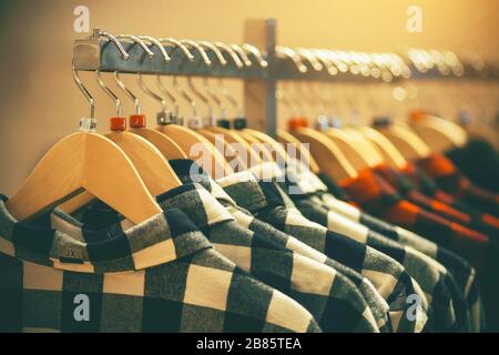 Nel negozio di abbigliamento, le camicie a scacchi di moda nei colori bianco e rosso di diverse dimensioni pendono sugli appendiabiti, illuminati dalla luce. Foto Stock