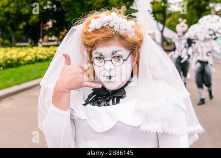 Una ragazza in un abito da sposa fa una telefonata con un gesto. Chiamami. Street performer in trucco e abbigliamento bianco al festival. Russia, Mosca - Foto Stock