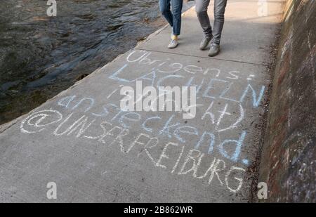 Friburgo, Germania. 20 Marzo 2020. Una coppia corre lungo il Dreisam. Con il gesso è dipinto su: 'A proposito, il sorriso è (non) contagioso @our Freiburg'. A causa della pandemia di Corona, la città di Friburgo ha emesso un coprifuoco per gruppi più grandi. Un cosiddetto divieto di ingresso nei luoghi pubblici è applicabile dal 21 marzo al 3 aprile Credit: Patrick Seeger/dpa/Alamy Live News Foto Stock