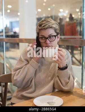 Donna che indossa un maglione avorio e occhiali neri, telefono a guglie, asciugando il naso e avendo una tazza di tè dopo lo shopping in un caffè Foto Stock