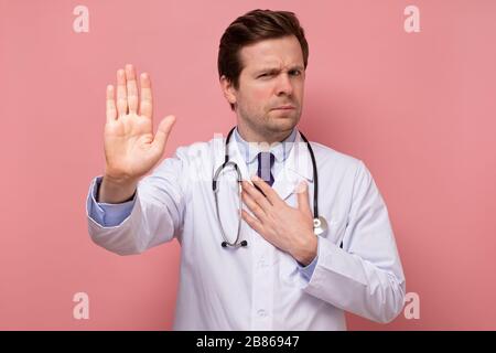 Giovane bell'uomo in cappotto bianco, interno o medico che mostra stop con la sua mano isolata su parete rosa. Foto Stock