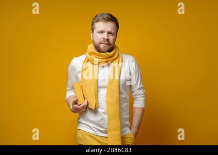 Allievo barbuto in una sciarpa gialla, lavorata a maglia, sta con un libro nelle mani e guarda con fiducia alla macchina fotografica. Concetto di istruzione. Foto Stock