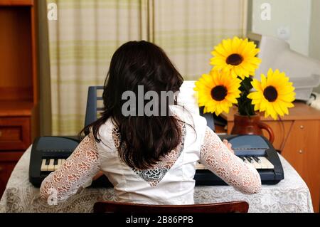 Bella donna brunetta in blusa bianca si siede e gioca sul sintetizzatore a casa. Interno sullo sfondo Foto Stock