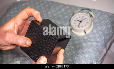 Un dito dell'uomo che preme il pulsante per accendere una macchina compatta, moderna, bianca, rumorosa, davanti ad una sveglia su un cuscino, pronta per andare a dormire. Foto Stock