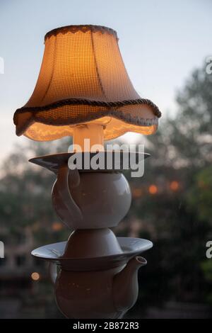 Lampada da tavolo in teiera vecchia tovana, piatti e tazze con paralume vintage sulla parte superiore. Lampada stile retro su sfondo sfocato attraverso la finestra. Foto Stock