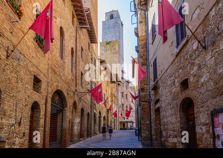 Due donne che camminano lungo via S. Matteo con bandiere rosse alle mura nel centro storico di San Gimignano, provincia di Siena, Italia. Foto Stock