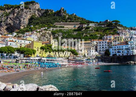 Vista sulla spiaggia con i suoi bagnanti dalla città di Amalfi dal molo con il mare, barche e case colorate sulle pendici della costiera amalfitana Foto Stock