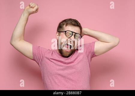 L'uomo attraente urla con la bocca aperta, chiudendo gli occhi e tirando le mani, su uno sfondo rosa. Foto Stock