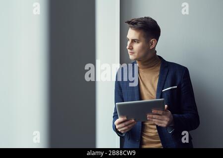 Giovane uomo d'affari in tuta in piedi e guardando attraverso la finestra mentre si utilizza il tablet digitale Foto Stock