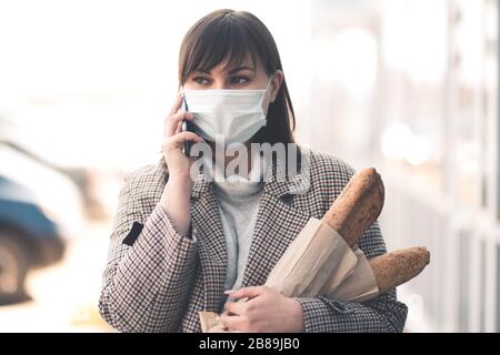 Giovane donna che indossa la maschera medica che parla al telefono tenendo il cibo camminare sulla strada all'aperto closeup. Guardando lontano. Concetto di virus. Distanza sociale. Foto Stock