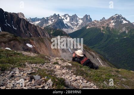 Un escursionista solitario che guarda attraverso il binocolo nelle Boundary Ranges delle Coast Mountains, sopra la Stikine River Valley, British Columbia, Canada. Foto Stock