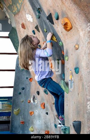 Una bambina attiva lavora per arrampicarsi su una parete di roccia interna al luogo di gioco di un bambino Foto Stock