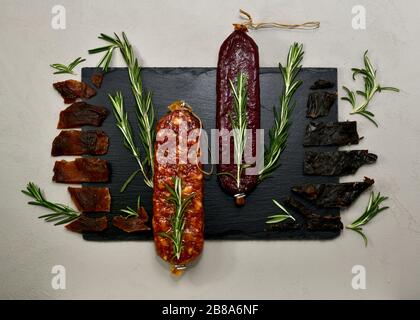 Prodotti di carne cruda affumicata e stagionata con rosmarino su fondo nero su fondo grigio in tonalità fresca. Foto Stock