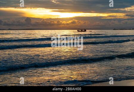 Tramonto lungo la spiaggia di Otres a Sihanoukville con la silhouette di persone che giocano nelle onde e l'ombra di una barca da pesca, Golfo della Thailandia, Cambogia. Foto Stock