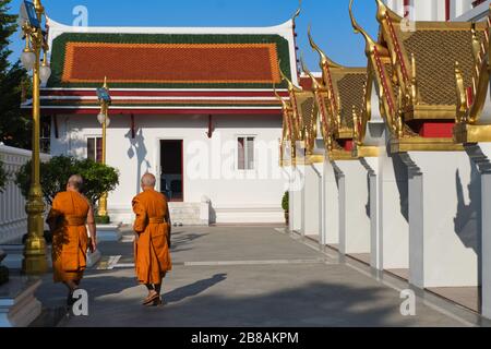 Due monaci buddisti con le rose arancioni camminano nei terreni di Wat Ratchanadta, la base dell'iconico edificio Lohaprasad sulla destra: Bangkok, Thailandia Foto Stock