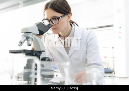 Giovane ricercatore contemporaneo in occhiali e whitecoat che guarda al microscopio mentre studia covid19 in laboratorio scientifico Foto Stock